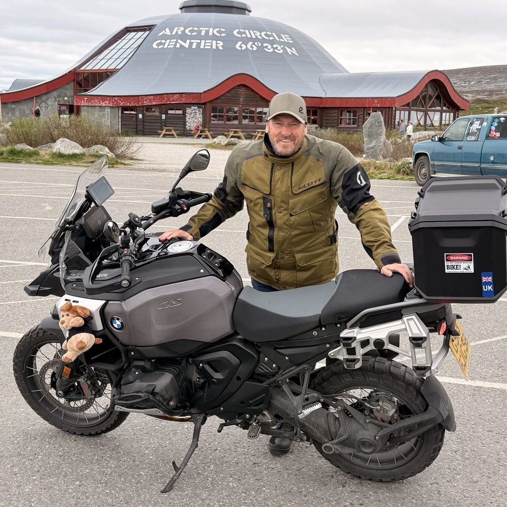James Slater in a cap and his olive riding jacket, leaning on his BMW GS in front of the Arctic Circle Center in Norway, its roof marked 66°33′N.