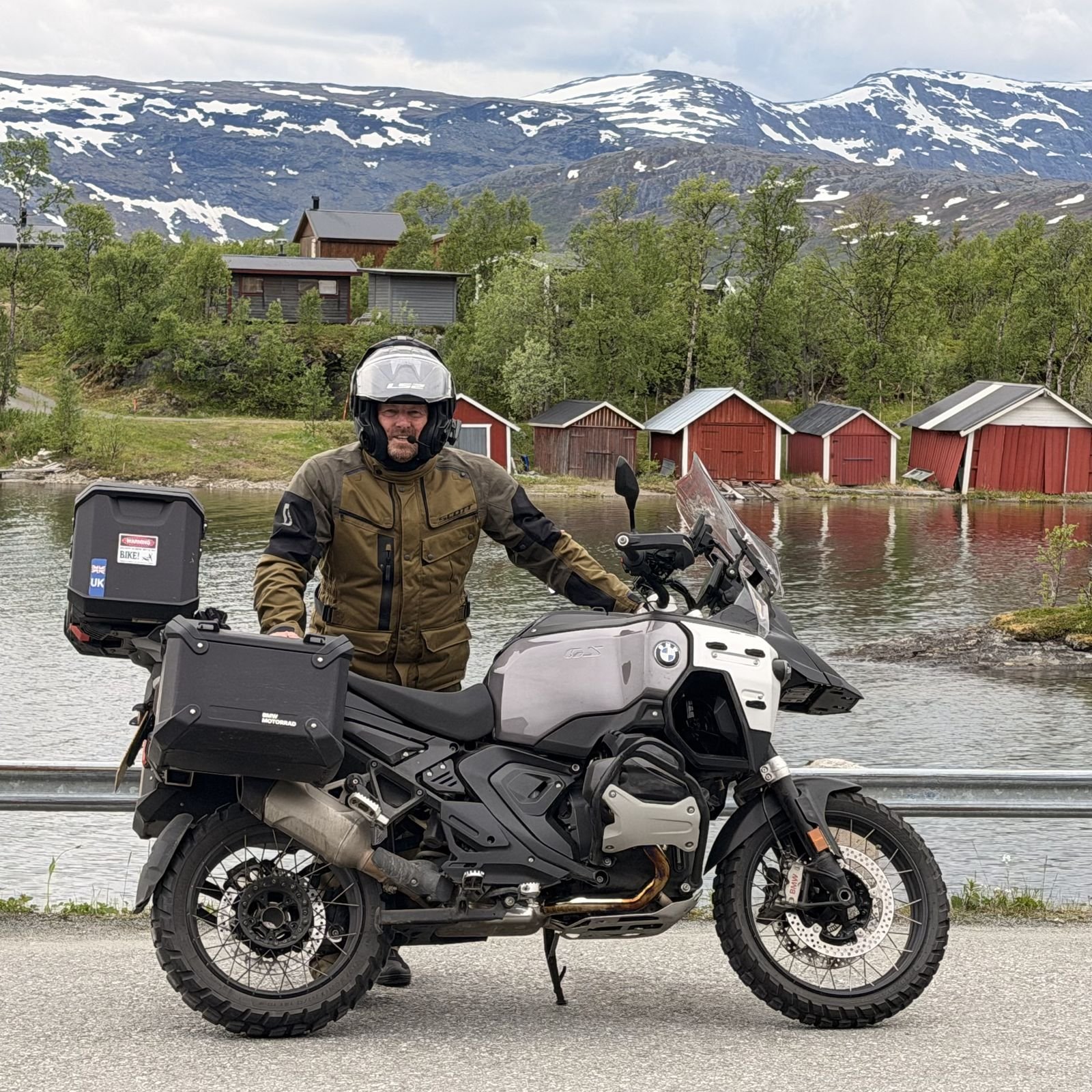 James Slater in olive adventure riding kit and a white modular helmet, standing behind his white and grey BMW GS with black panniers and a top box, red wooden boathouses on a lake and snow-capped mountains behind him in Norway.